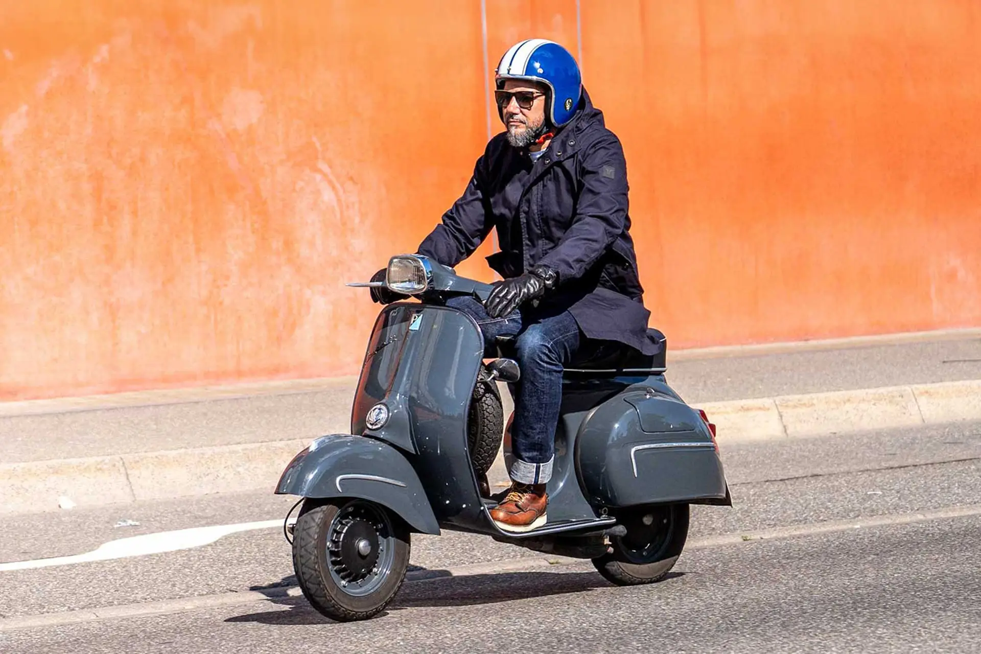 Vespa-Fahrer trägt den grünen Urban Mobility Parka von Caduff vor einer orangefarbenen Wand, perfekt für den urbanen Alltag mit Schutz bis 45 km/h. Vespa rider wearing the green Urban Mobility Parka by Caduff in front of an orange wall, perfect for urban daily commutes with protection up to 45 km/h.